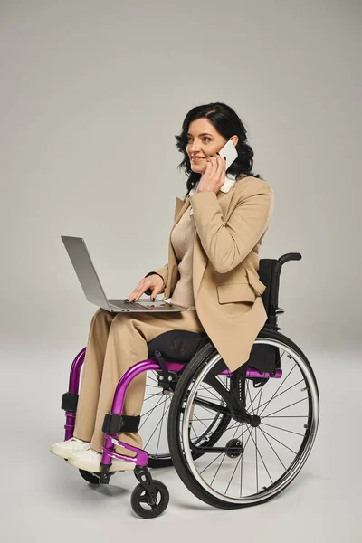 Mujer alegre con discapacidad de movilidad en silla de ruedas con su portátil y hablando por teléfono - foto de stock