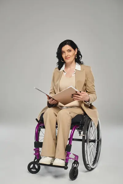 Hermosa mujer morena con discapacidad sentada en silla de ruedas con papeleo y mirando a la cámara - foto de stock