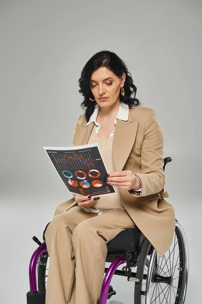 Mujer morena con discapacidad de movilidad en traje pastel mirando gráficos mientras está en silla de ruedas - foto de stock