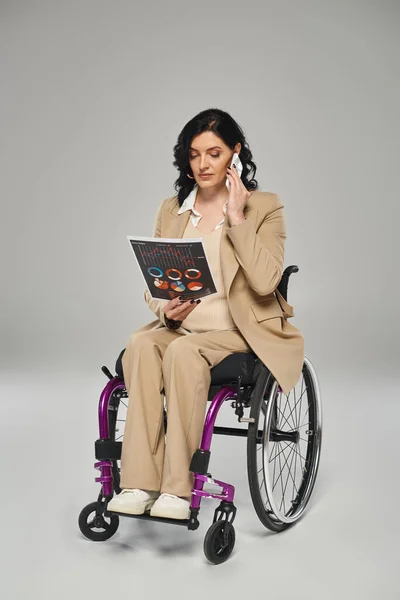 Hermosa mujer discapacitada en traje elegante discutir gráficos por teléfono mientras está sentado en silla de ruedas - foto de stock