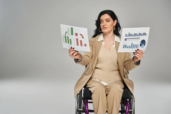 Séduisante femme handicapée avec les cheveux ondulés en costume chic en fauteuil roulant en regardant les graphiques dans ses mains — Photo de stock