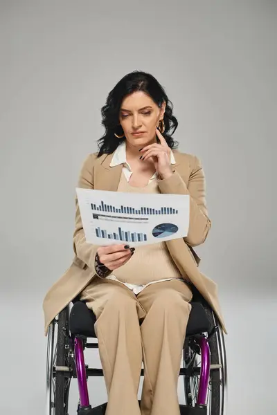 Mujer discapacitada enfocada con cabello ondulado en traje pastel sentada en silla de ruedas y mirando gráficos - foto de stock