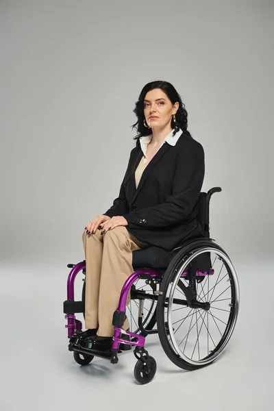 Mulher atraente com deficiência em blazer preto sentado em cadeira de rodas e olhando para a câmera — Fotografia de Stock