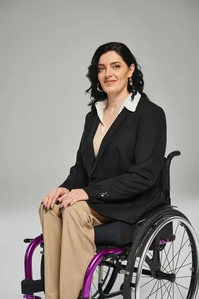 Alegre morena con discapacidad en chaqueta negra sentada en silla de ruedas y sonriendo a la cámara - foto de stock