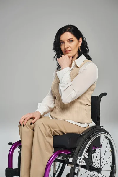Atractiva mujer morena con discapacidad en elegante atuendo pastel en silla de ruedas mirando a la cámara - foto de stock