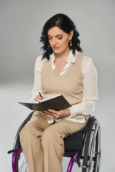Hermosa mujer morena con discapacidad en atuendo pastel en silla de ruedas mirando su cuaderno - foto de stock