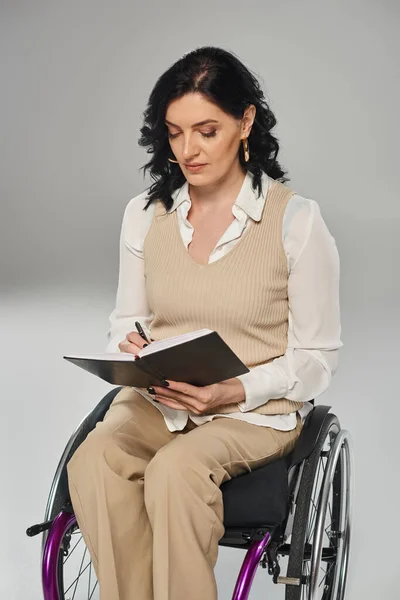 Beautiful brunette woman with disability in pastel attire in wheelchair looking at her notebook — Stock Photo