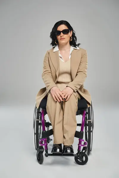 Atractiva mujer discapacitada con el pelo ondulado sentado en silla de ruedas en gafas de sol y mirando a la cámara - foto de stock