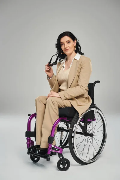 Schöne selbstbewusste Frau mit Behinderung im Rollstuhl, die mit Sonnenbrille in der Hand in die Kamera blickt — Stockfoto