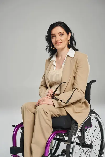 Mujer atractiva alegre con discapacidad en silla de ruedas sosteniendo gafas de sol y mirando a la cámara - foto de stock