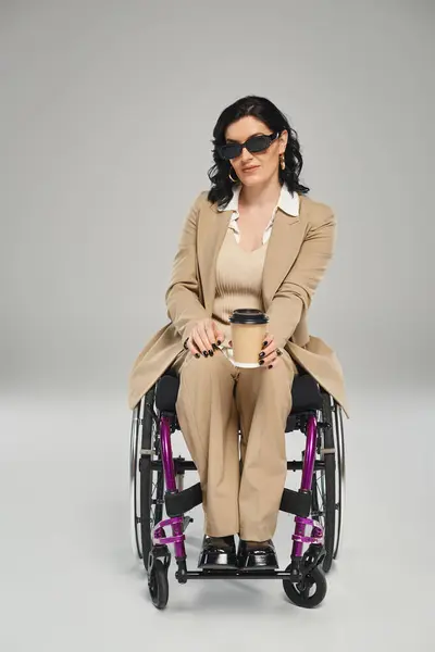 Belle femme handicapée en pastel tenue élégante avec des lunettes de soleil en fauteuil roulant tenant café — Photo de stock