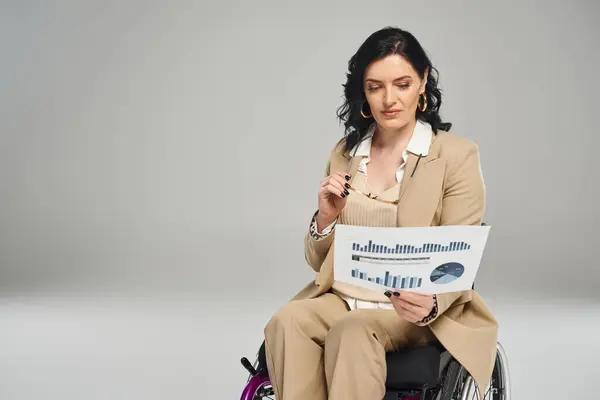Mujer guapa con discapacidad en silla de ruedas sosteniendo sus gafas y mirando sus gráficos - foto de stock