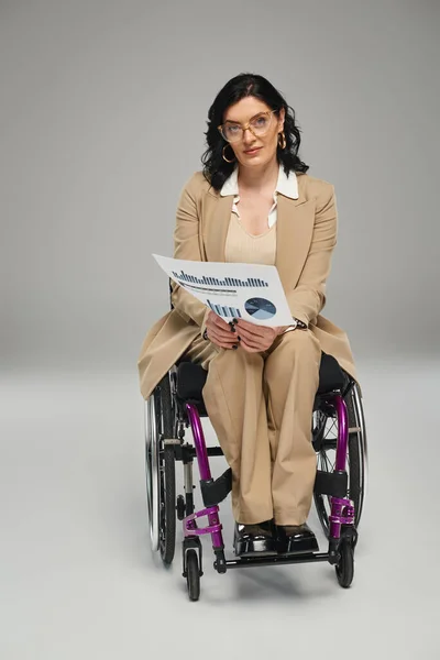 Mujer discapacitada enfocada con gafas y cabello ondulado en su silla de ruedas con gráficos mirando a la cámara - foto de stock