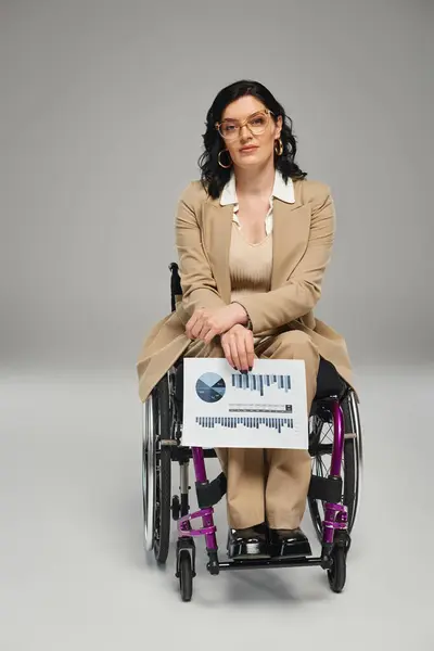 Attrayant femme handicapée avec des lunettes en fauteuil roulant regardant la caméra avec des graphiques à la main — Photo de stock