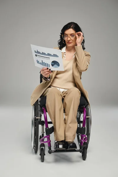 Jolie femme brune handicapée avec des lunettes de travail sur les graphiques en fauteuil roulant — Photo de stock