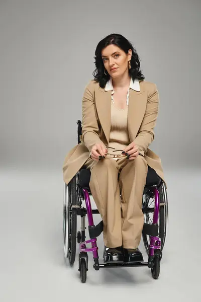 Mujer segura de buen aspecto en traje de pastel en silla de ruedas sosteniendo gafas y mirando a la cámara - foto de stock