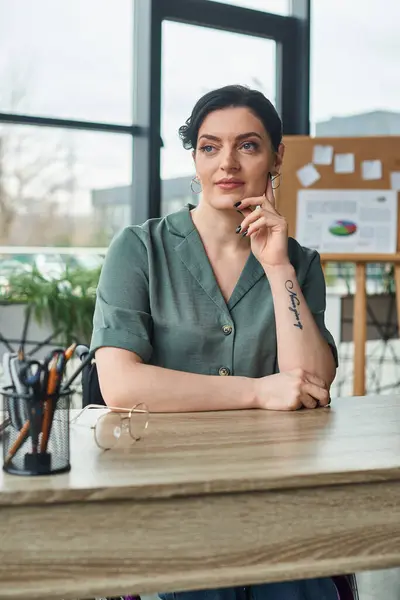 Attractive confident businesswoman in casual attire sitting at table and looking away in office — Stock Photo