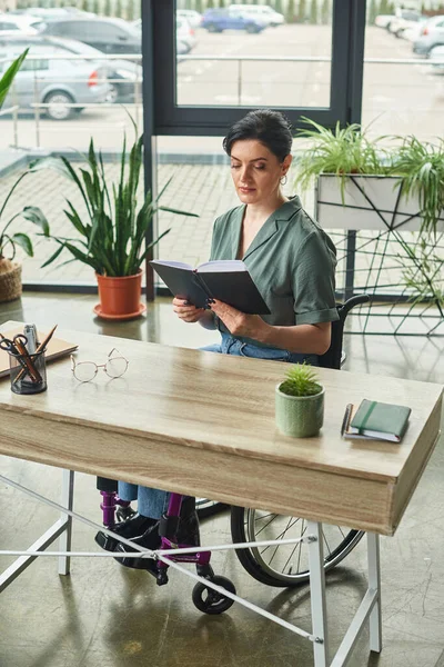 Mulher morena confiante em roupa casual em cadeira de rodas trabalhando com seu caderno no escritório — Fotografia de Stock