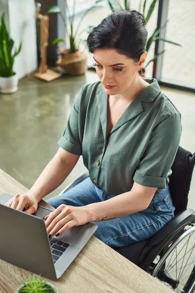 Привлекательная уверенная деловая женщина с татуировкой в инвалидной коляске, работающая за своим ноутбуком в офисе — стоковое фото