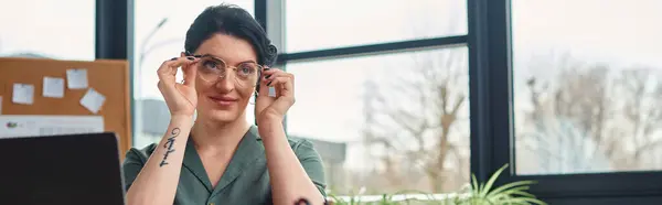Atractiva mujer discapacitada en traje casual con tatuaje y gafas mirando hacia otro lado mientras está en la oficina - foto de stock