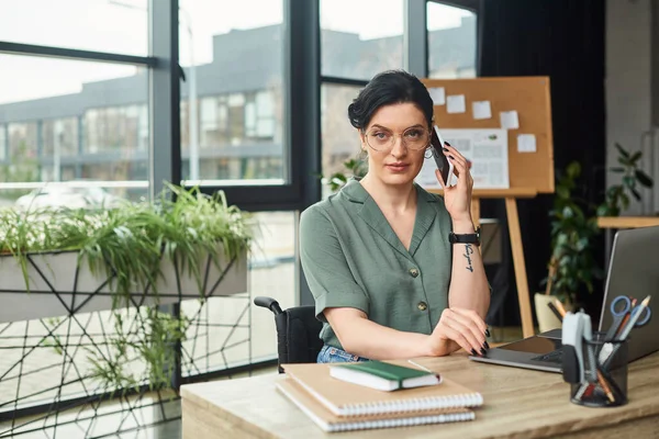 Задумчивая, красивая женщина-инвалид в повседневной одежде в очках разговаривает по телефону во время работы — стоковое фото