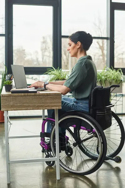 Attrayant femme handicapée en tenue décontractée assis en fauteuil roulant travaillant sur son ordinateur portable au bureau — Photo de stock