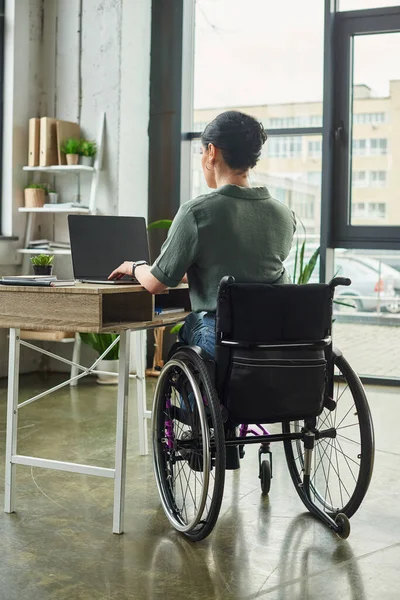 Femme handicapée brune dédiée en tenue décontractée assis en fauteuil roulant et travaillant dur au bureau — Photo de stock