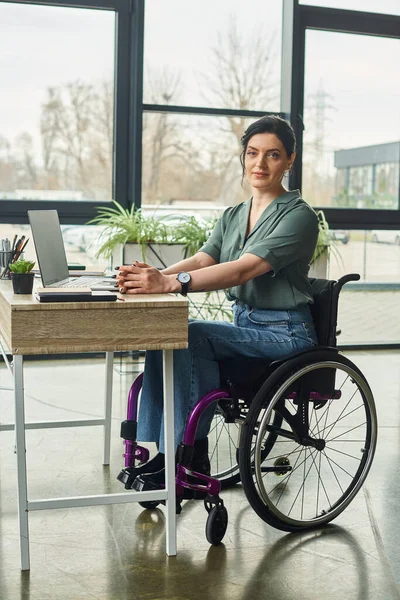 Joyeuse femme d'affaires handicapée en tenue décontractée en fauteuil roulant regardant la caméra tout en travaillant dur — Photo de stock