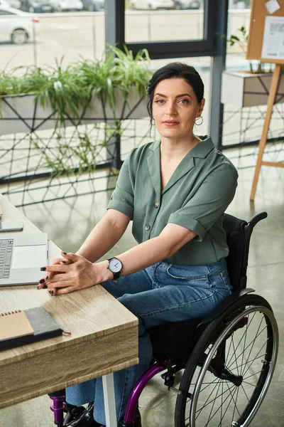 Красивая, уверенная в себе женщина-инвалид в повседневной одежде в инвалидном кресле, смотрит в камеру на работе — стоковое фото