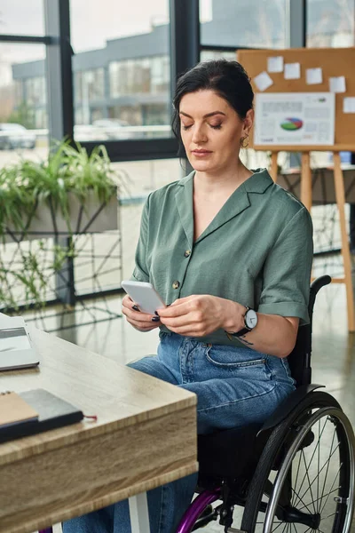 Hermosa mujer de negocios dedicada con discapacidad en silla de ruedas mirando su teléfono inteligente en la oficina - foto de stock