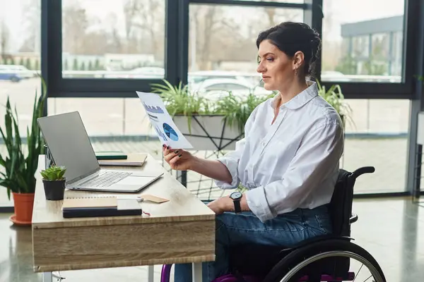 Donna d'affari dedicata con disabilità in sedia a rotelle guardando la sua grafica mentre in ufficio — Foto stock