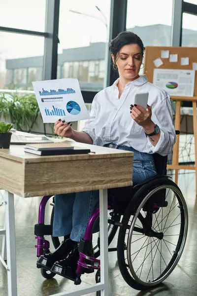 Atractiva mujer discapacitada vestida con estilo en silla de ruedas mirando el teléfono mientras trabaja con gráficos - foto de stock