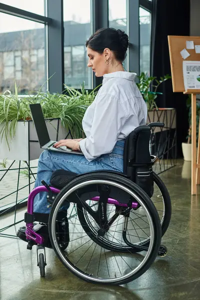 Преданная деловая женщина с ограниченными возможностями в городской одежде в инвалидной коляске работает на своем ноутбуке в офисе — стоковое фото