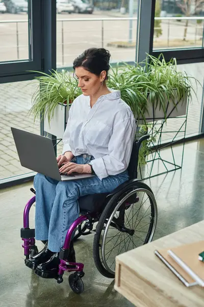 Внимательная деловая женщина-инвалид в повседневной одежде в инвалидной коляске работает за ноутбуком во время работы в офисе — стоковое фото
