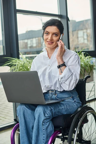 Femme handicapée gaie en fauteuil roulant parlant par téléphone tenant ordinateur portable et regardant la caméra — Photo de stock