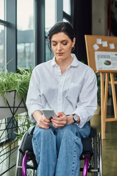 Хорошая трудолюбивая женщина с инвалидностью в инвалидной коляске, глядя на свой смартфон в офисе — стоковое фото