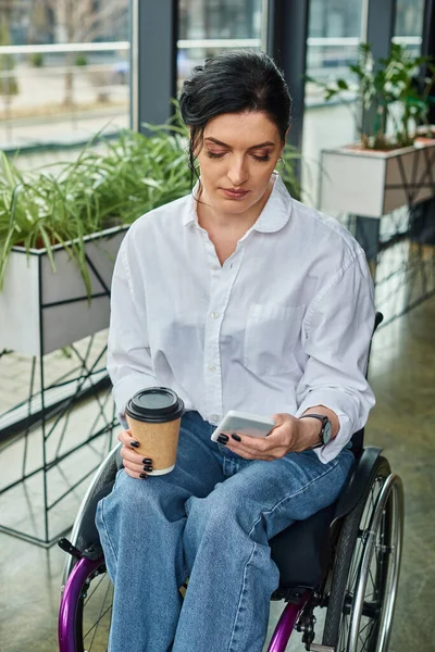 Atractiva morena con discapacidad en silla de ruedas sosteniendo café y mirando su teléfono - foto de stock
