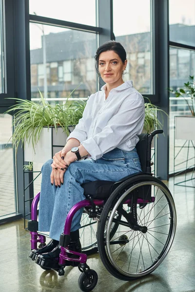 Весьма привлекательная деловая женщина-инвалид в инвалидном кресле, счастливо улыбающаяся в камеру во время работы в офисе — стоковое фото