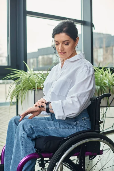 Bella donna disabile in abbigliamento chic seduto sulla sedia a rotelle e guardando il suo orologio da polso — Foto stock