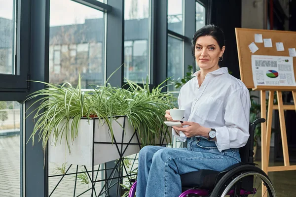 Jolly beautiful disabled woman in business attire in wheelchair holding coffee and smiling at camera — Stock Photo