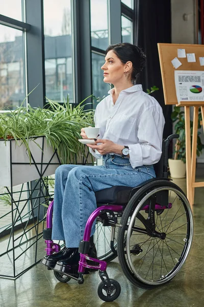 Привлекательная преданная деловая женщина с ограниченными возможностями в инвалидном кресле держит чашку кофе и смотрит в сторону — стоковое фото
