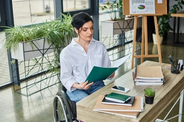 Уверенная в себе предпринимательница-инвалид в инвалидной коляске просматривает свои документы на своем рабочем месте в офисе — стоковое фото