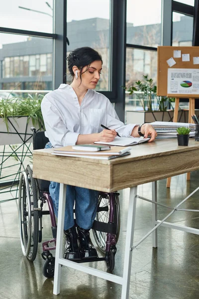 Хорошо выглядящая трудолюбивая деловая женщина с инвалидностью в инвалидном кресле делает заметки за столом — стоковое фото