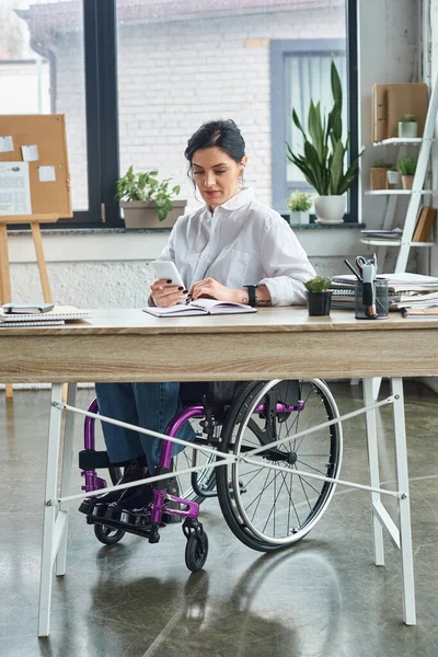 Bella donna d'affari bruna con disabilità in sedia a rotelle guardando il suo telefono in ufficio — Foto stock