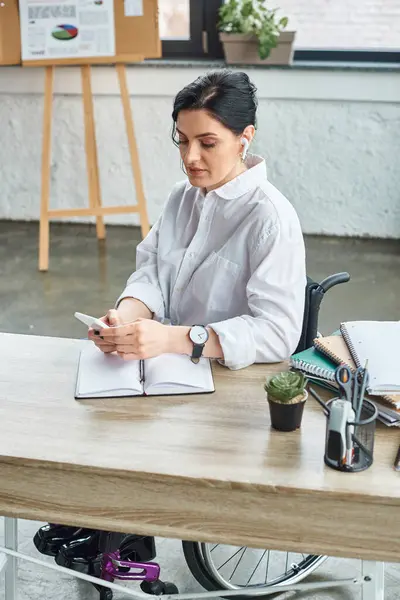 Привлекательная женщина-инвалид в стильной одежде с наушниками, смотрящая на свой телефон в офисе — стоковое фото