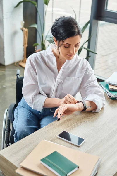 Gut aussehende, behinderte Geschäftsfrau im Rollstuhl überprüft im Büro ihre Armbanduhr — Stockfoto