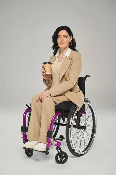 Atractiva mujer discapacitada en traje de pastel en silla de ruedas sosteniendo café y mirando a la cámara - foto de stock
