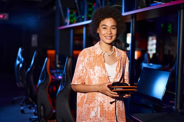 Happy african american woman wearing casual clothing holding headphones in cybersport club — Stock Photo