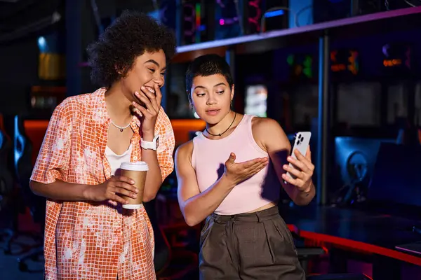 Две стильные межрасовые женщины делятся моментом радости и общения за чашкой кофе, глядя в телефон — стоковое фото