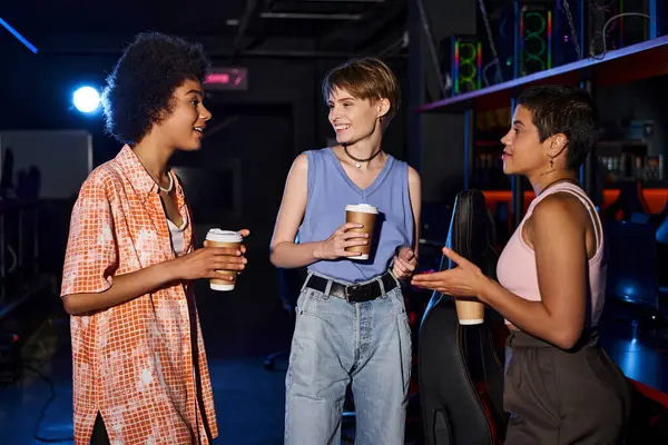 Un grupo diverso de mujeres elegantes sosteniendo tazas de café hablando y reuniéndose en una habitación oscura - foto de stock